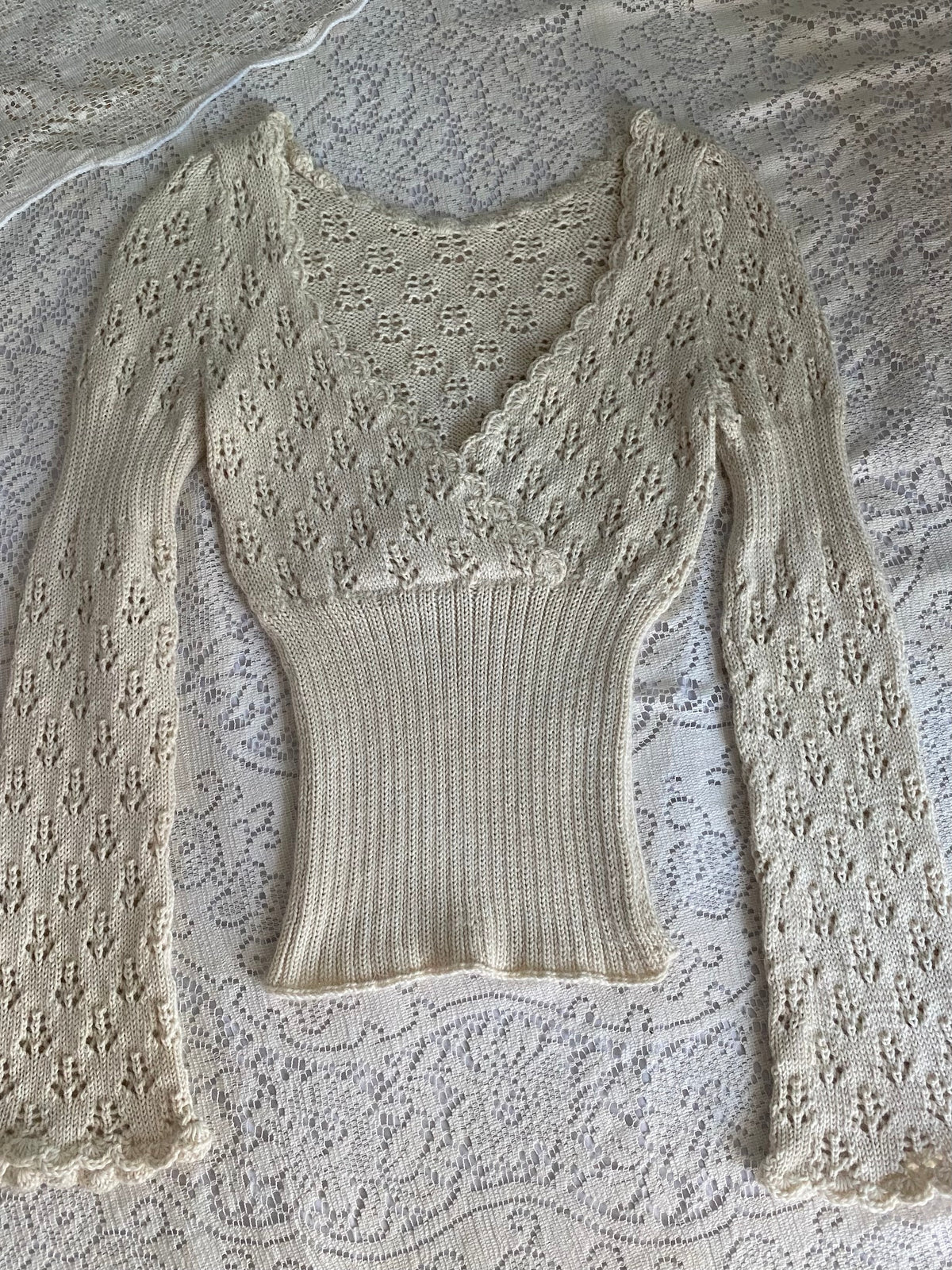 A cream-colored, long-sleeve knit top with a lace overlay and ribbed bodice, laid flat on a white lace background.